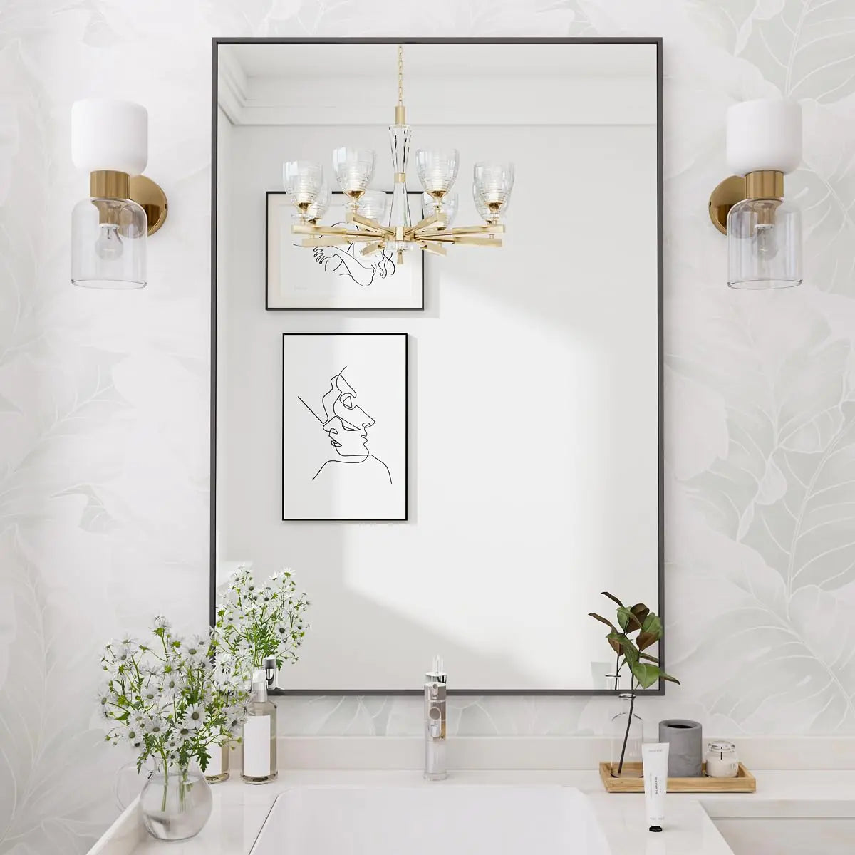 Modern rectangular black-framed bathroom mirror above a white vanity with gold wall sconces, floral wallpaper, minimalist line art, and fresh white flowers in a vase.