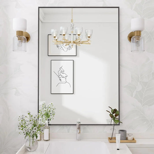 Modern rectangular black-framed bathroom mirror above a white vanity with gold wall sconces, floral wallpaper, minimalist line art, and fresh white flowers in a vase.
